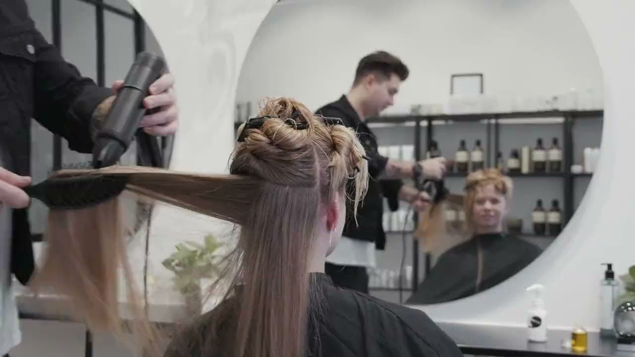 A stylist blow-drying a client's long hair in front of a round mirror